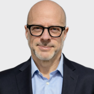 A bald man with glasses, wearing a dark suit jacket over a light blue shirt, is smiling slightly at the camera against a plain light background. EFESO Management Consultants